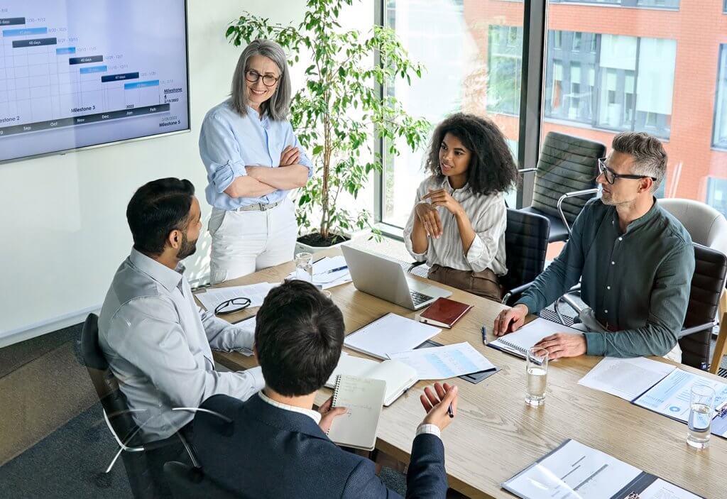 A woman with gray hair, arms crossed, stands and leads a meeting in a glass-walled conference room, addressing four colleagues seated around a wooden table. A wall-mounted screen behind her displays a project timeline with milestones. The seated group includes a woman with curly hair near a laptop, two men in business casual attire, and a man in a suit writing in a notebook in the foreground. Water glasses, notepads, and documents are spread across the table.